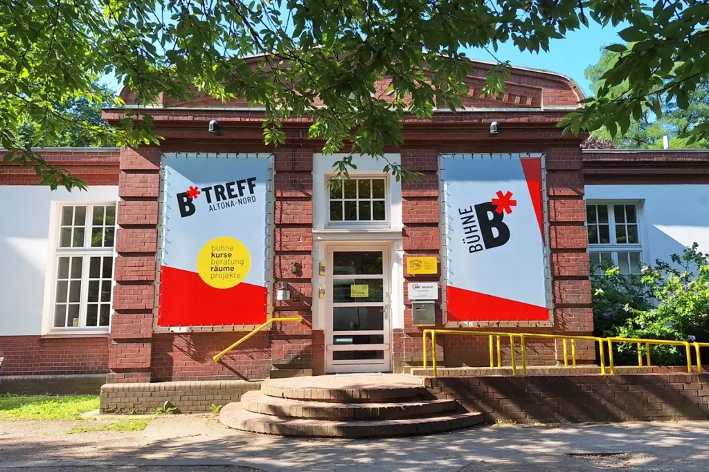 Außenansicht B-Treff Altona Nord. Backstein-Gebäude mit gelben Geländern und großen Bannern, die "BTreff Altona-Nord" und "Bühne B" zeigen.